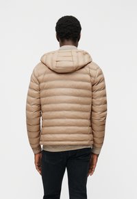 Άνδρας που φοράει μπεζ καπιτονέ μπουφάν με κουκούλα και σκούρο παντελόνι, φαίνεται από πίσω πάνω σε απλό λευκό φόντο.