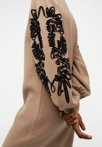 Robe beige avec de longues manches bouffantes ornées de broderies noires complexes. Texture de tissu lisse avec des motifs décoratifs et des accents de nœuds.