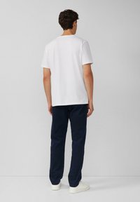 Weißes Baumwoll-T-Shirt mit kurzen Ärmeln, kombiniert mit navyblauen Hosen und weißen Schuhen, von hinten vor einem neutralen Hintergrund betrachtet.