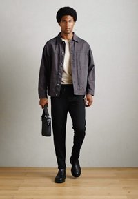Gray jacket over a cream shirt, black pants, and black boots. Holding a black bag. Wood floor and light gray background.