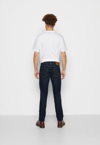 Hombre de pie frente a una pared blanca, vistiendo una camisa blanca de manga corta, jeans azul oscuro con bolsillos traseros y zapatos marrones.