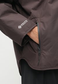 Rjava vetroodporna jakna z gladko teksturo, stranskim žepom na zadrgo in logotipom na rokavu. Nastavljiv rob z ukrivljenim dizajnom.