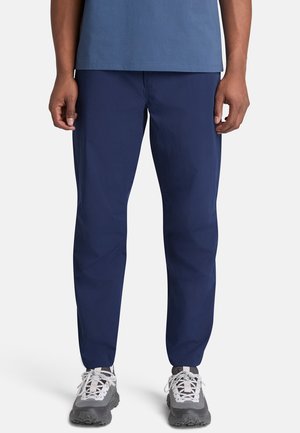 Homme portant un pantalon slim bleu foncé, des chaussures de sport grises et blanches, et une chemise bleue, debout devant un fond clair uni.