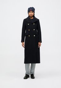 Dobbeltbrystet uldfrakke i marineblå med guldknapper, slagkrave og sidelommer, parret med en mørk strikket beanie og jeans.