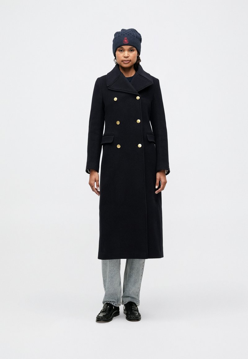 Dobbeltbrystet uldfrakke i marineblå med guldknapper, slagkrave og sidelommer, parret med en mørk strikket beanie og jeans.