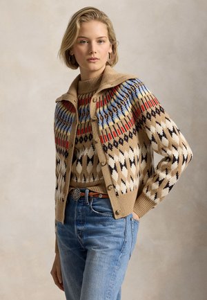 Femme portant un cardigan beige à motifs sur un pull assorti, un jean bleu avec une ceinture décorative et des mocassins marron, debout les mains dans les poches.