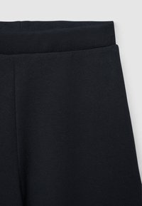 Primo piano di una cintura elastica nera su pantaloni morbidi a gamba dritta con cucitura visibile lungo la parte anteriore.