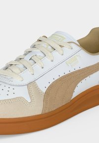 Λευκό και μπεζ εσωτερικό αθλητικό παπούτσι Puma με καουτσούκ σόλα, κρεμ κορδόνια και λογότυπο στο πλάι και στη γλώσσα.