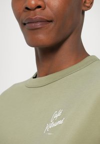 Sudadera verde oliva con cuello redondo y logotipo bordado "Café Kitsuné" en blanco. Textura suave, detalle de costura visible.