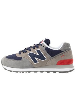 Grauer und navyfarbener New Balance 574 Sneaker mit rotem Fersenakzent, weißer Sohle und strukturiertem Wildleder- und Mesh-Obermaterial, Seitenansicht.