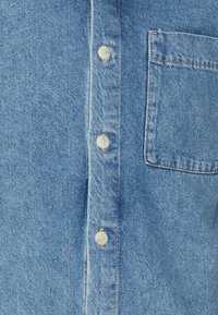Camicia in denim azzurro chiaro con chiusura a bottoni, caratterizzata da bottoni bianchi e una tasca sul petto a sinistra. Il tessuto ha una finitura strutturata.