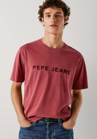 Camiseta roja de algodón de manga corta con cuello redondo. Tiene la inscripción "PEPE JEANS" en letras negras en la parte delantera. Corte informal.
