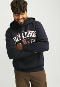 Navy blue hooded sweatshirt with front graphic text and a kangaroo pocket, featuring ribbed cuffs and hem, paired with brown cargo pants.