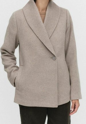 Béžový vlněný kabát s širokými klopami, zapínáním na jeden knoflík a bočními kapsami, nošený přes světlé tričko a tmavé kalhoty.