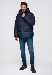 Granatowa kurtka puchowa z kapturem, z zamkiem błyskawicznym i zapięciem na guzik, z pikowaną dekoracją. Połączona z niebieskimi dżinsami i czarnymi sznurowanymi botkami.