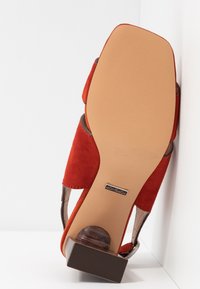 Červené semišové sandály na podpatku s hnědým texturovaným blokovým podpatkem. Podešev je béžová s hladkým povrchem. Vlastnosti zahrnují otevřené strany a bezpečný pás na kotníku.