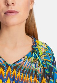 Colorful blouse featuring a geometric pattern in blue, yellow, and green. The V-neck design has gathered accents near the neckline.