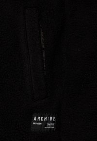 Svart fleecejacka med en strukturerad yta, dold ficka och ett framträdande märke med "ARCHIVE ONLY & SONS"-logotyp.