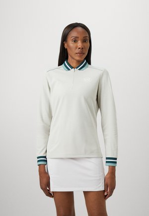 Vrouw in een wit langemouwen polo shirt met groene gestreepte kraag en manchetten, en een witte rok tegen een effen achtergrond.