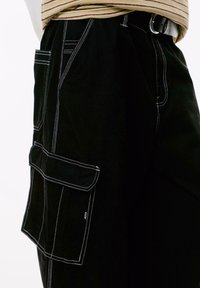 Pantalons cargo noirs avec une grande poche latérale et des coutures blanches contrastées, portés avec un haut striped beige rentré et une ceinture noire.