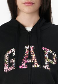 Black zip-up hoodie featuring a floral "GAP" logo in pink, white, and green, with textured embroidery and a drawstring hood.