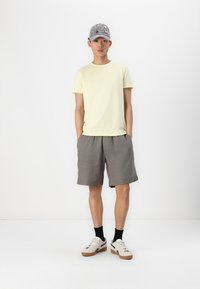 Lichtgele crew-neck t-shirt, grijze shorts, witte sneakers met tan accenten en een grijze pet met multicultureel geborduurde details.