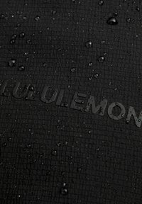 Zbliżenie czarnego materiału ripstop z kroplami wody osiadającymi na powierzchni oraz ledwo widocznym wytłoczonym napisem "LULULEMON".