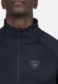 Maglione in pile nero con zip e colletto alto, caratterizzato da un motivo testurizzato, logo Rossignol in grigio tenue sulla zona sinistra del petto.