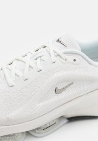 La scarpa sportiva bianca presenta una tomaia in mesh testurizzato, chiusura con lacci, il logo Nike in argento e un'unità di ammortizzazione Zoom Air visibile.