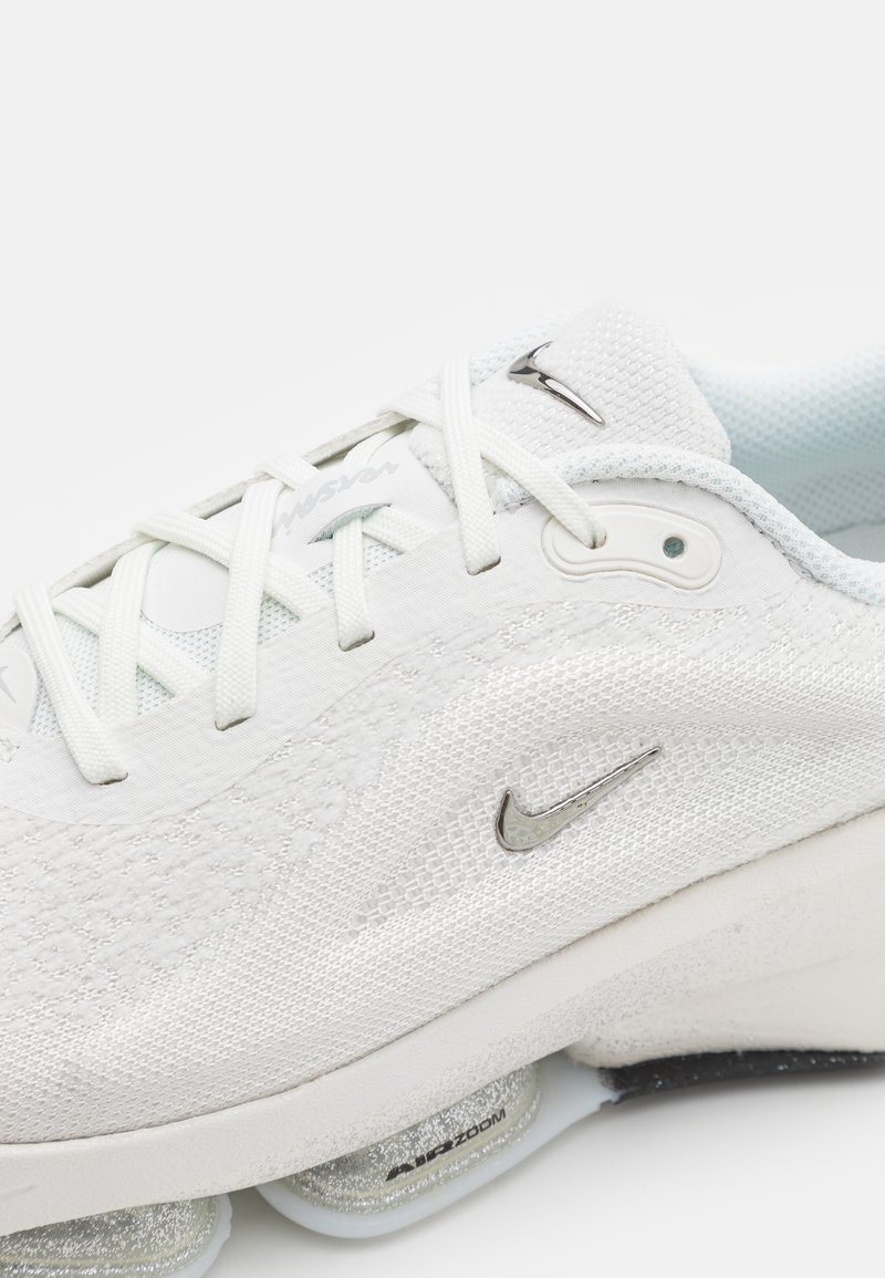 Zapatilla deportiva blanca con parte superior de malla texturizada, cierre de cordones, logo de Nike en plata y una unidad de amortiguación Zoom Air visible.