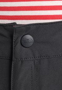 Svarta tygbyxor har en rund svart knapp med "The North Face"-logotyp. Bakgrunden visar röd och vit randig tyg.