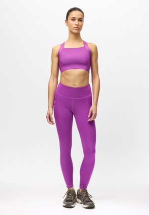 Vrouw staand, draagt een felpaarse sportbeha en leggings met grijze hardloopschoenen op een effen witte achtergrond.