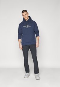 Hoodie bleu marine avec une poche avant et du texte blanc, associé à un pantalon sombre et des baskets gris clair. Présente une coupe décontractée et un design à capuche.