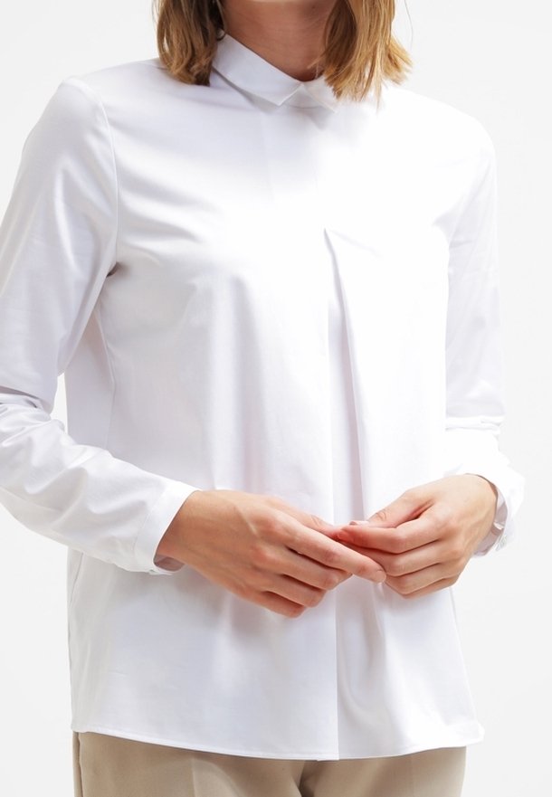 Personne portant une chemise blanche à manches longues boutonnée avec un col montant, tenant ses mains ensemble au niveau de la taille.