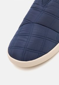Navy blue quilted fabric sneaker with a rounded toe, beige sole, and contrasting blue stitching, featuring a slip-on design.