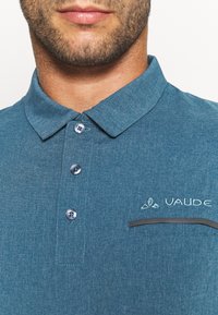 Gros plan sur le bas du visage et le cou d'un homme portant un polo bleu à col avec trois boutons et une petite poche sur la poitrine.