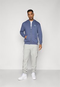 Blå hættetrøje med lynlås og logo, parret med lysegrå cargo-sweatpants og hvide sneakers. Enkel design med lommer foran.