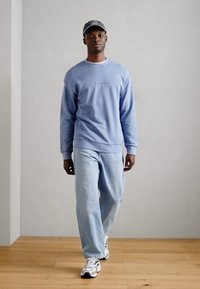 Sudadera azul claro con puños y dobladillo acanalados, combinada con jeans azul claro de corte relajado y zapatillas blancas. Gorra gris con detalle de logo.