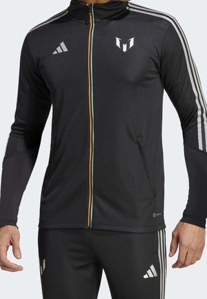 Mężczyzna ubrany w czarny dres Adidas z białymi paskami na rękawach i nogawkach, z zamkiem błyskawicznym z pomarańczowo-niebieską lamówką oraz logo na piersi i spodniach.