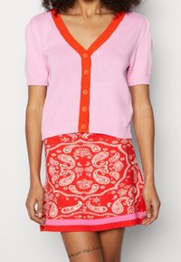Rosa kortärmad cardigan med djup V-ringning och orange knappslå, ihopparad med en röd paisleymönstrad kjol med rosa kant.