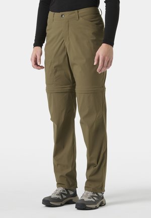 Mężczyzna w oliwkowych spodniach trekkingowych z odpinanymi nogawkami i szarych butach terenowych, stojący na jasnym, jednolitym tle.