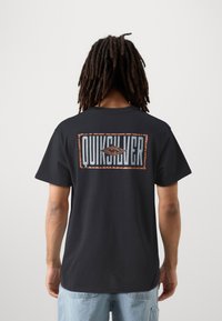 Black cotton t-shirt featuring a large "Quiksilver" logo on the back with a textured graphic and a rectangular outline.