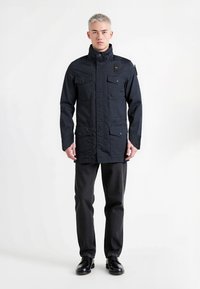 Granatowa kurtka z wysokim kołnierzem, czterema przednimi kieszeniami i zatrzaskami. Materiał wydaje się trwały; noszona na ciemnych spodniach i butach.