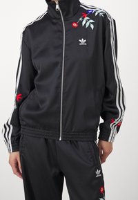 Svart idrottsträningjacka med hög krage, dragkedja, vita ränder och blommig brodyr i röd och blå på axlarna.