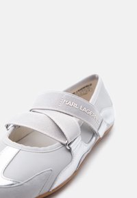 Λευκό παπούτσι χωρίς κορδόνια με φαρδιές ελαστικές λουρίδες που διασταυρώνονται, με την επωνυμία "Karl Lagerfeld" και ανοιχτό καφέ σόλα, εμφανιζόμενο σε ελαφρώς κεκλιμένη γωνία.