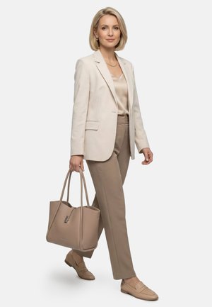 Blonde vrouw in beige blazer en broek, lopend, met een grote taupekleurige handtas, met bijpassende loafers en gouden sieraden.