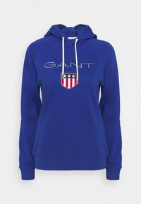 Modrá mikina s kapucí na stahování, na které je nápis "GANT" v světle šedé barvě a design s štítem, hvězdami a pruhy. Měkký materiál.