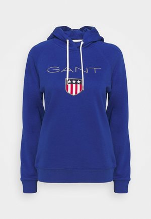 Sudadera azul con capucha con cordón, que presenta la palabra "GANT" en gris claro y un diseño de escudo con estrellas y franjas. Material suave.