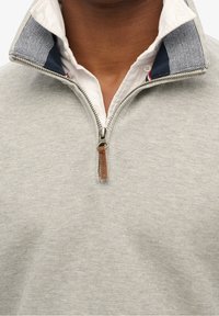 Pullover grigio chiaro con zip frontale e tirazip in pelle. Presenta una texture a maglia e un colletto a righe blu sotto.