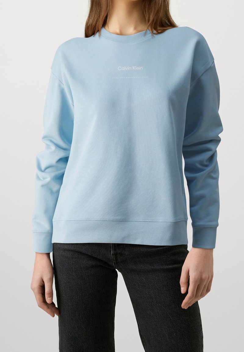Femme portant un sweat-shirt Calvin Klein bleu clair à manches longues et un pantalon noir, debout devant un fond blanc uni.